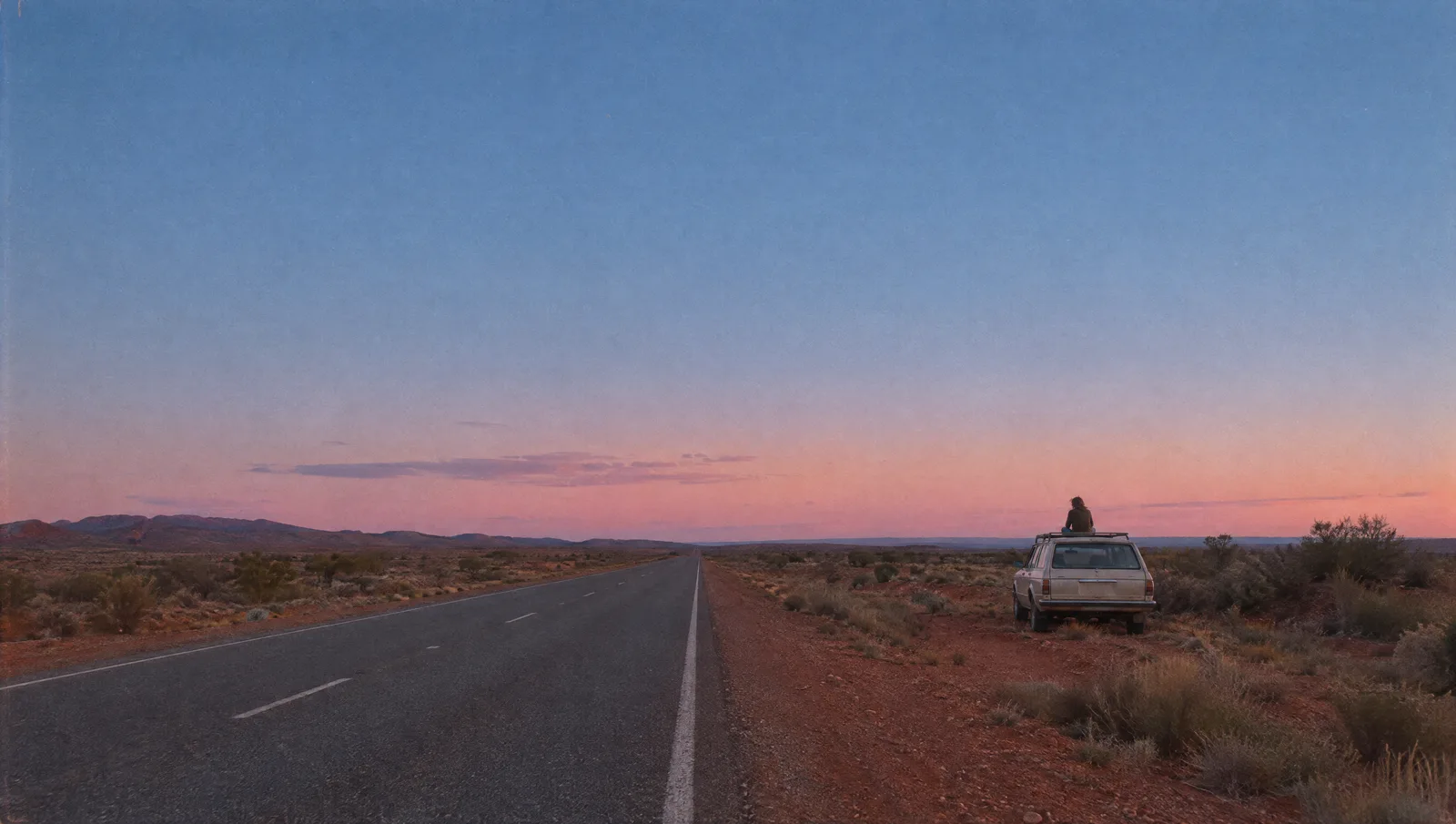 A station wagon parked on a red desert road at dusk, a figure sitting on its roof